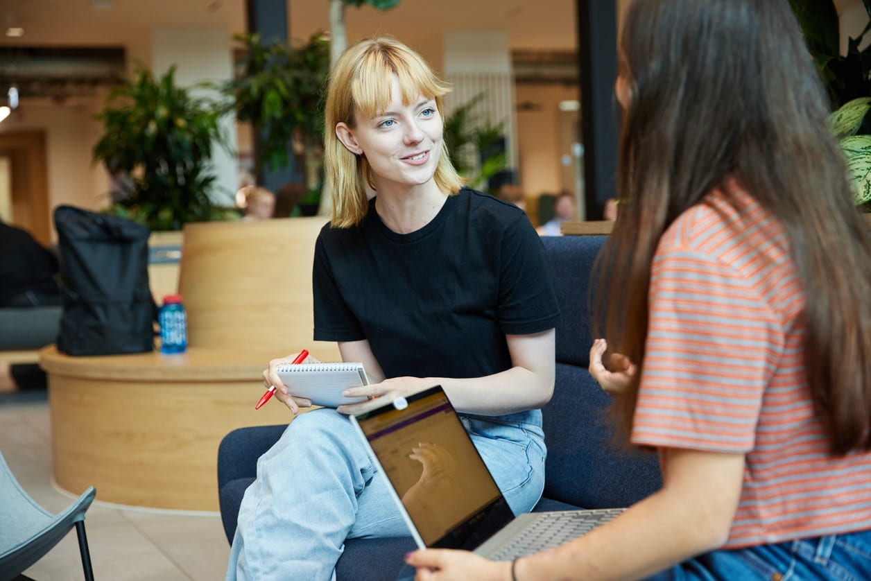 female student sitting down with laptop open, smiling at another female student in the foreground with her back to the camera in an open cafe environment.