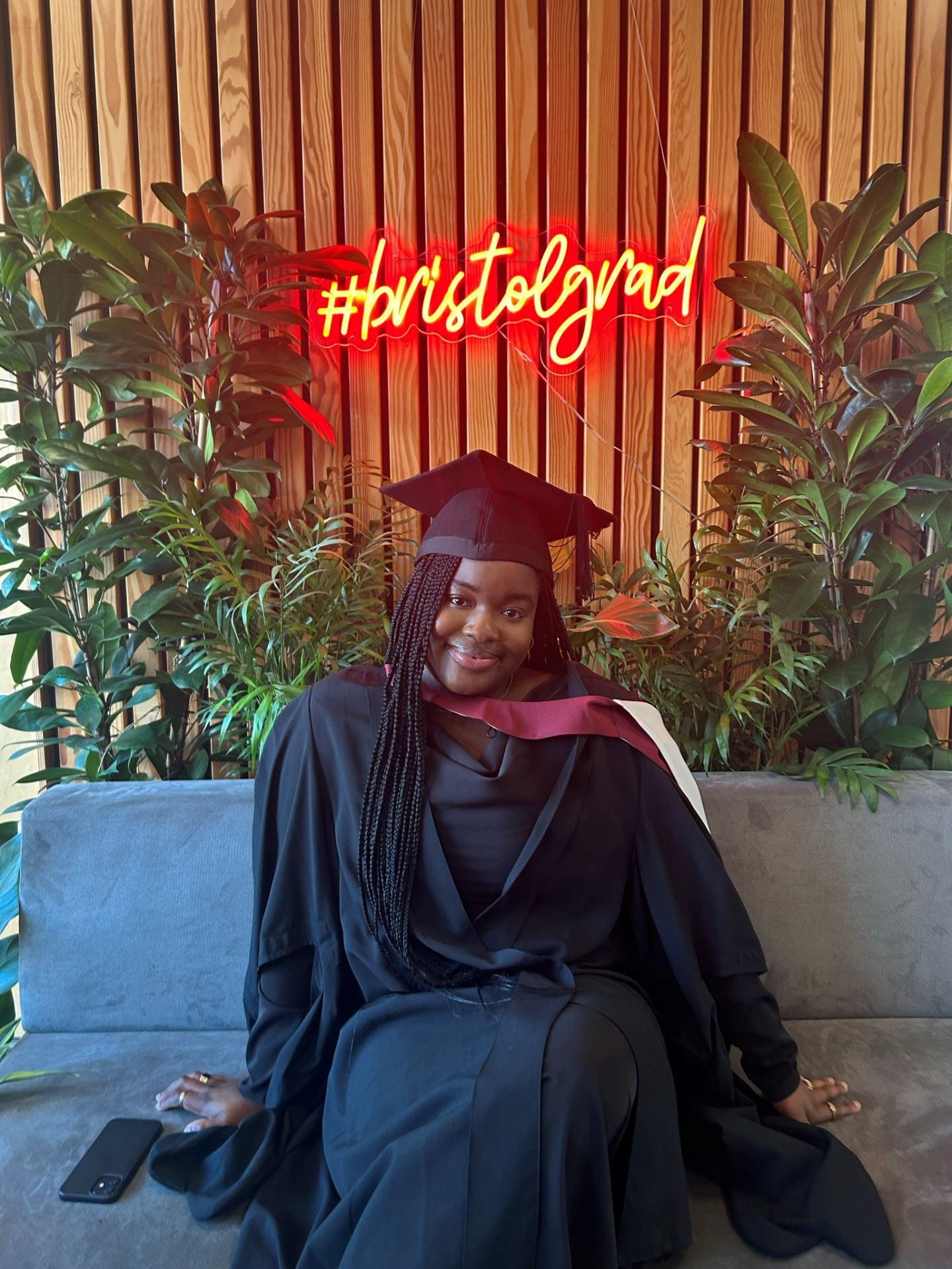 law student, Oladayo Ige, wearing a black graduation gown and cap, sitting in front a wooden panneled wall covered in plants with a neon sign saying #bristolgrad.