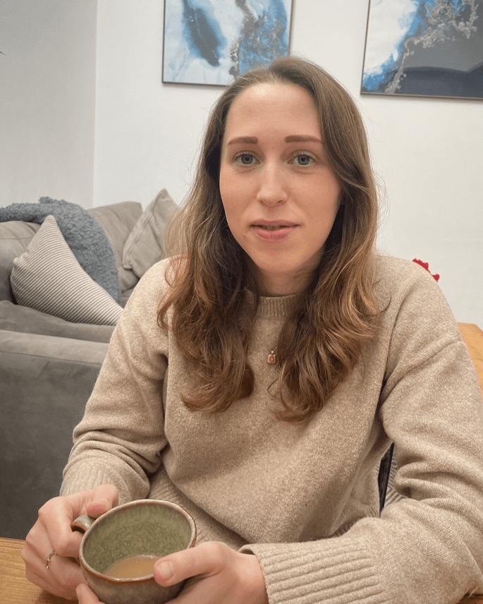 Image of Alex Walker, UNiverisyt of Bristol Law Clinic Solicitor and Teaching Associate, wearing a beige jumper, sat down in fron tof a sofa with a mug in her hands.
